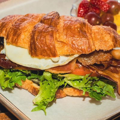 A BLT sandwich made with a croissant style bread, including bacon, leafy lettuce, with a fried egg for breakfast nutrition